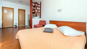 Quarto de hotel com cama, uma cadeira vermelha, estante de madeira e duas portas.