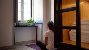 A person sitting on a purple mat in a room with a window, a plant on a shelf, and a wooden cabinet. - GiG Capsule Hostel (Mafra)