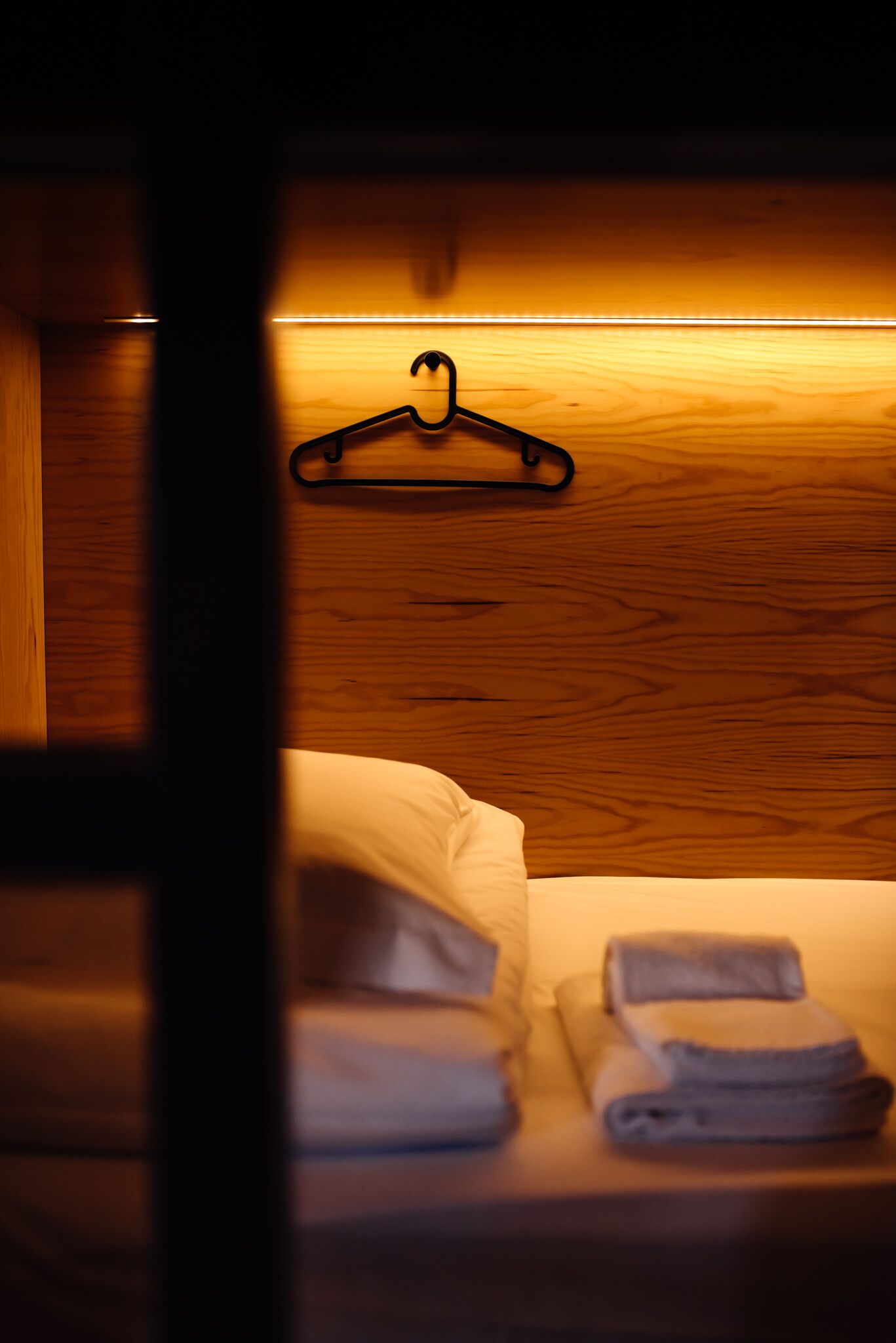 A neatly made bed with folded towels and a black hanger on a wooden wall.