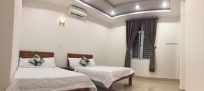 A hotel room with two single beds, a ceiling fan, an air conditioning unit, a window with curtains, and a staircase visible through the window.