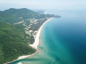 An aerial view of a coastal area with a beach, forested hills, and a clear blue sea.
