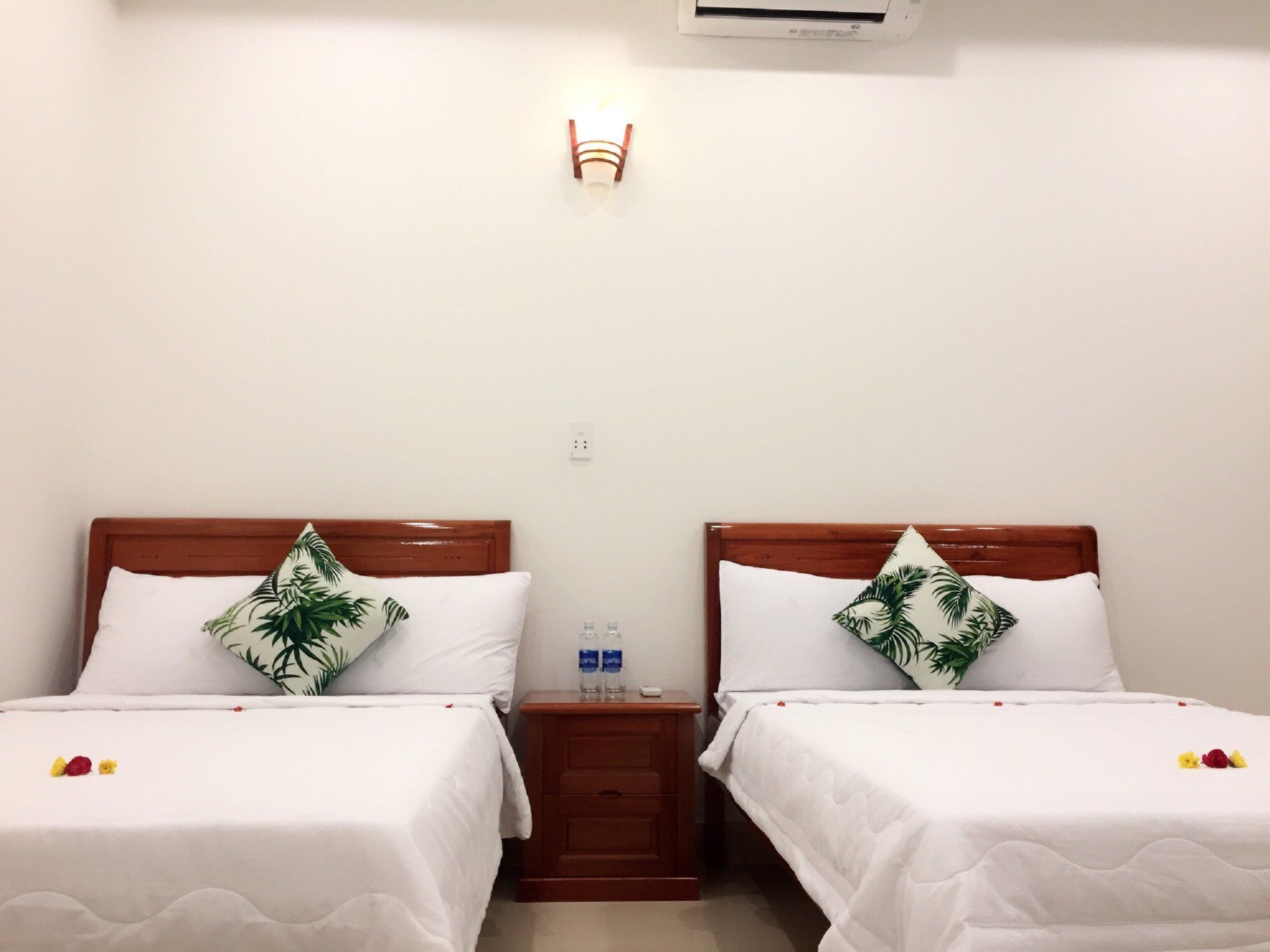 Quarto de hotel com duas camas de solteiro, cada uma com cabeceira de madeira e roupa de cama branca. Há uma mesinha de cabeceira entre as camas, com uma garrafa e um pequeno objeto sobre ela. Um candeeiro fixado na parede está acima da mesinha.