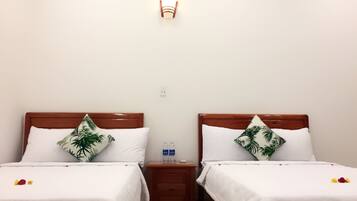 Quarto de hotel com duas camas de solteiro, cada uma com cabeceira de madeira e roupa de cama branca. Há uma mesinha de cabeceira entre as camas, com uma garrafa e um pequeno objeto sobre ela. Um candeeiro fixado na parede está acima da mesinha.