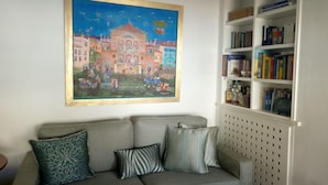 A living room with a grey sofa, patterned cushions, a bookshelf with books, and a colorful painting of a building. - Luxury Flat with Amazing Lycabetus View (Athens)