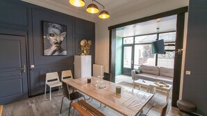 Un espace repas moderne avec une table en bois, des chaises et un banc. On y trouve une grande fenêtre avec des stores, un canapé et un téléviseur fixé au mur.