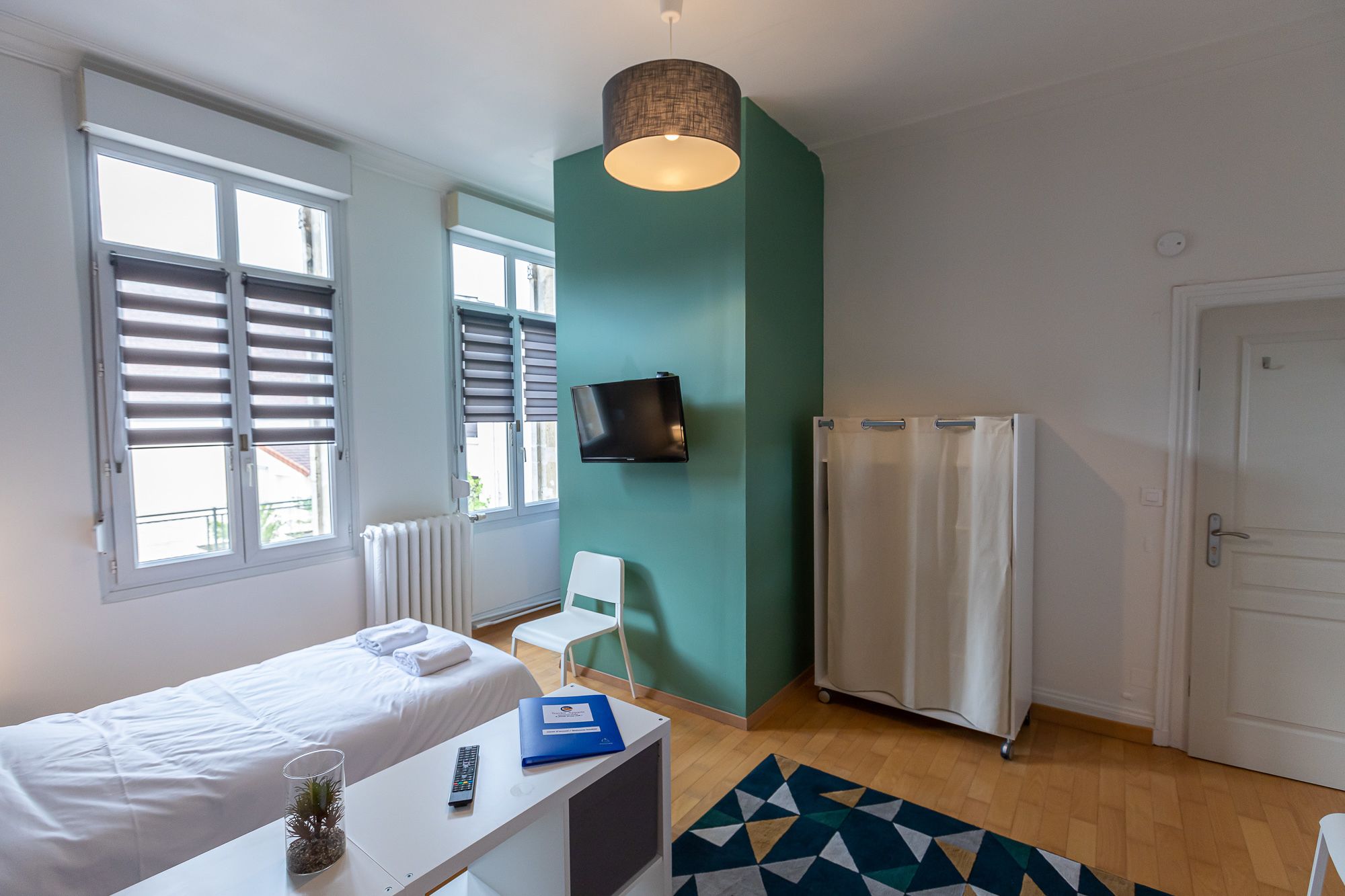 A hotel room with a bed, a desk with a remote control and a folder, a TV on the wall, a window with blinds, a radiator, and a shower curtain.
