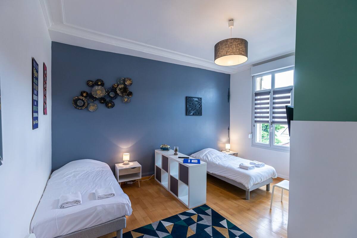A bedroom with two beds, a bedside table with a lamp, a small shelf, and a geometric rug.