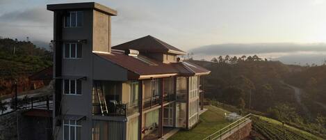A modern multi-story house with a balcony, large windows, and a view of a hilly landscape.