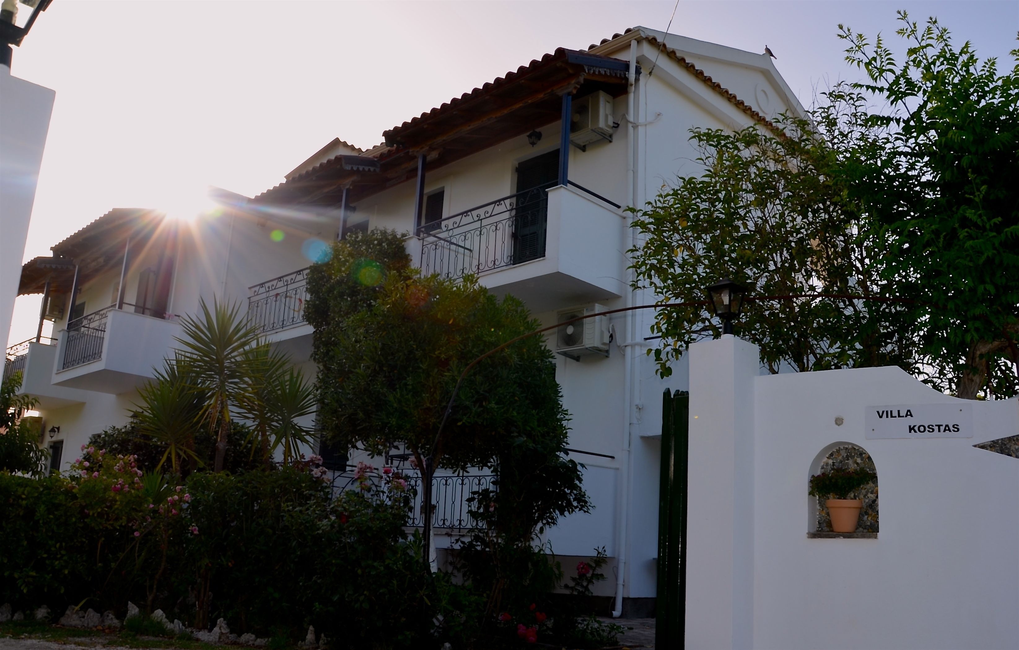 A two-story building with a balcony, surrounded by greenery and a sign reading "VILLA KOSTAS".