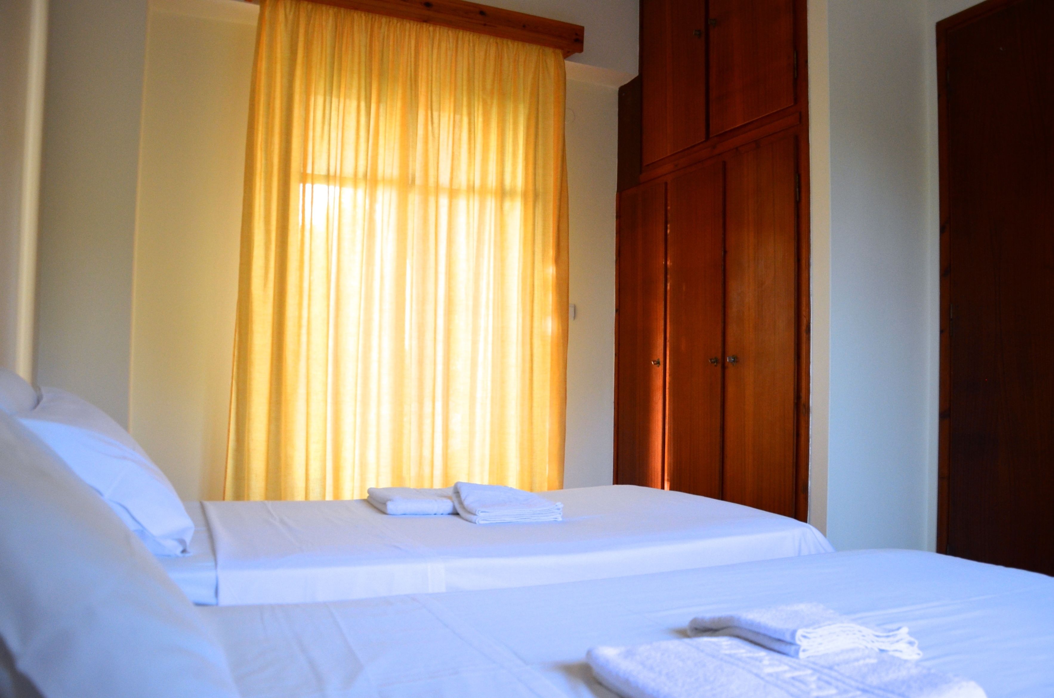 A hotel room with a bed, a wooden wardrobe, and a window with yellow curtains.