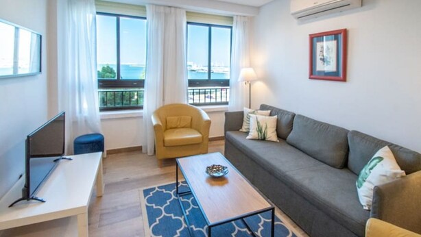A modern living room with a sofa, a small table, a chair, and a TV. There are windows with a view of the sea. - Apartamento Albatro´s suite (Cádiz)