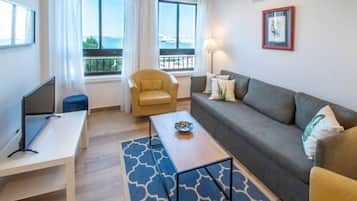 A modern living room with a sofa, a small table, a chair, and a TV. There are windows with a view of the sea.