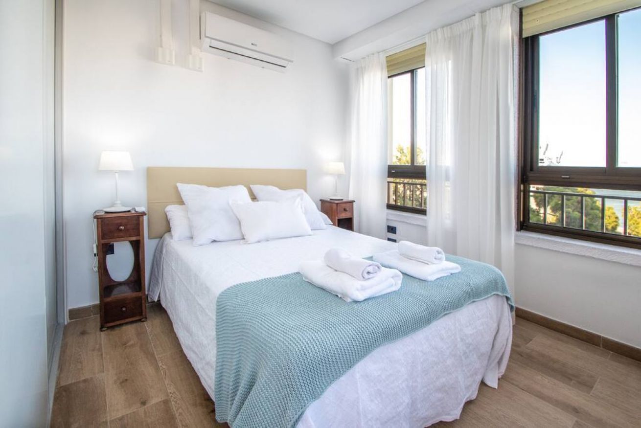 A neatly made bed with white linens and a teal blanket, a bedside table with a lamp, and a window with a view of trees.