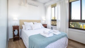 A neatly made bed with white linens and a teal blanket, a bedside table with a lamp, and a window with a view of trees. - Apartamento Albatro´s suite (Cádiz)