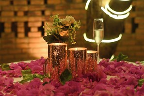 Three copper candle holders with flowers and a glass of champagne on a table covered with rose petals. - Saj By The Lake (Murbad)