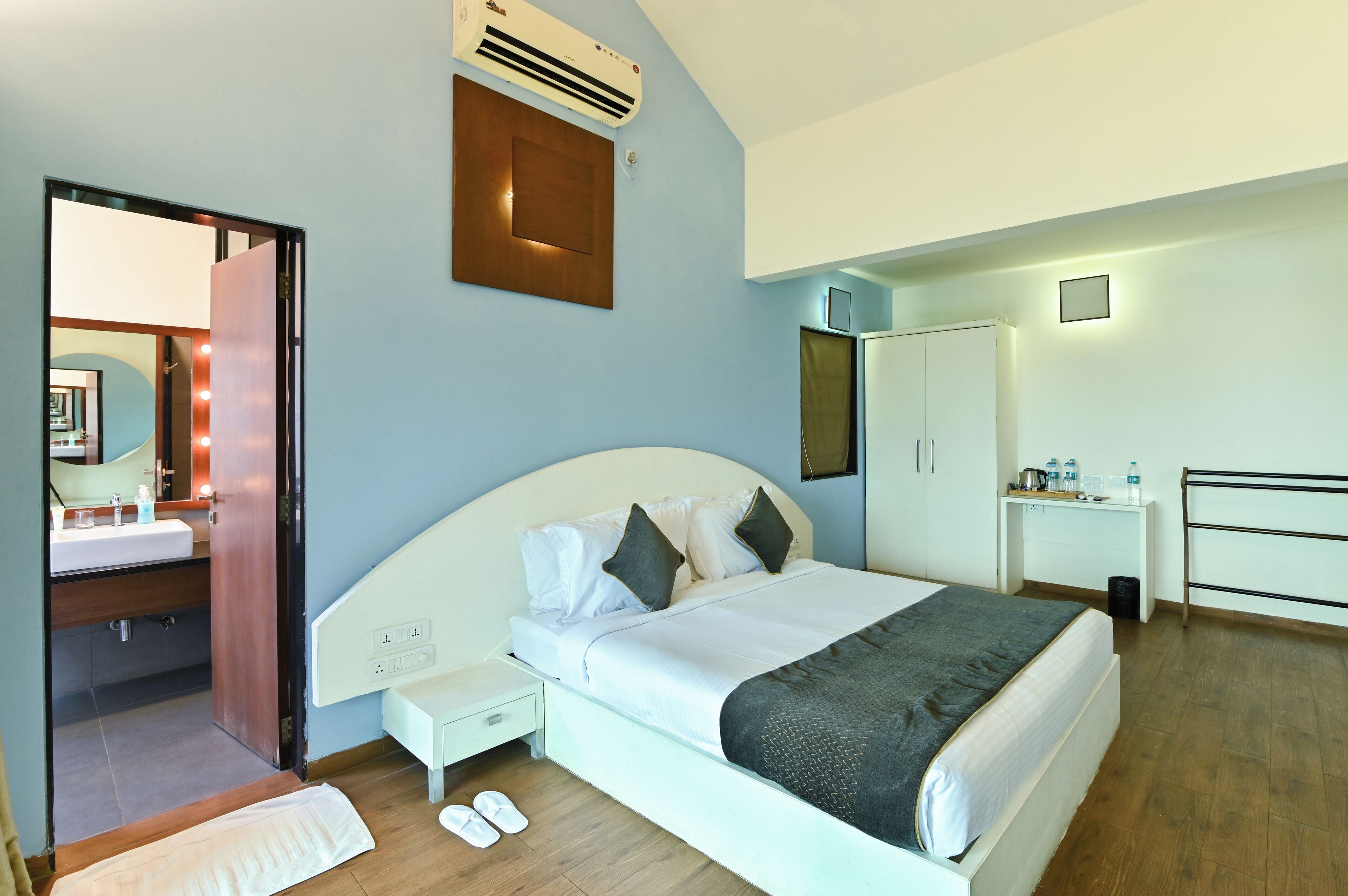 A modern hotel room with a large bed, bedside table, and a small desk with a chair. There is a wardrobe and a mirror on the wall.