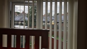 Une pièce dotée d’une fenêtre avec des stores verticaux, un garde-corps rouge et une vue sur les maisons voisines, qui sont équipées de panneaux solaires.