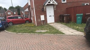 Une rue résidentielle bordée de maisons, avec une voiture rouge garée devant l’une d’elles et une poubelle verte portant l’inscription « Tottenham Hotspur Football Club ».