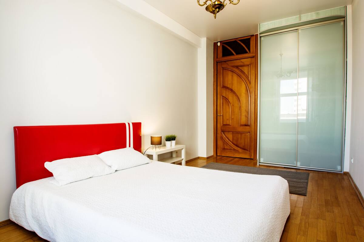 A bedroom with a red headboard, a white bedspread, a wooden door, and a sliding glass closet.