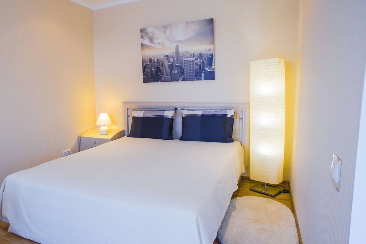 A hotel room with a bed, a nightstand with a lamp, a wall-mounted painting, and a large floor lamp.