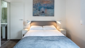 Un dormitorio con cama, dos lámparas de noche, una mesita de noche, un cuadro en la pared y un baño visible a través de una puerta abierta.