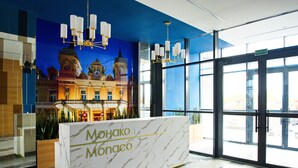 Ein Empfangsschalter mit der Beschriftung "MOHAKO MONACO", einer Kristallleuchtre und einem Wandbild eines Gebäudes.