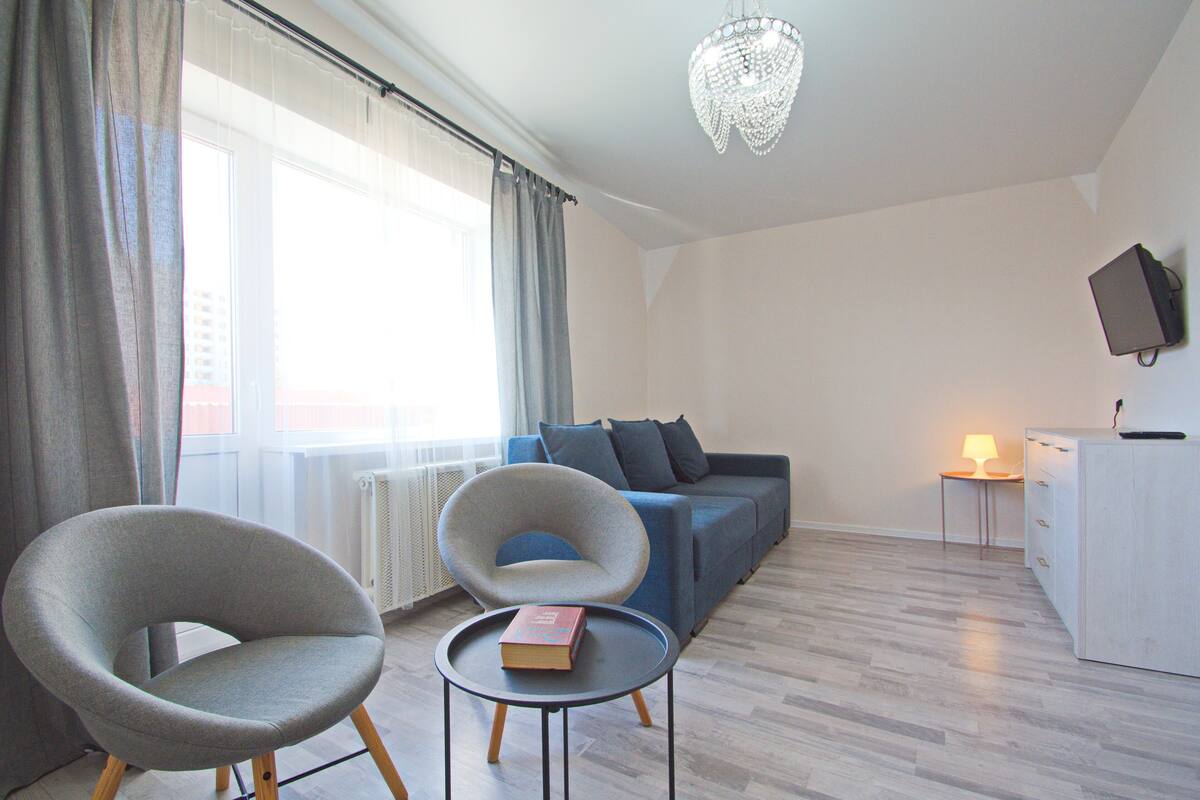 Una sala moderna con un sofá, dos sillones, una mesita con un libro, una lámpara y un televisor sobre un mueble.