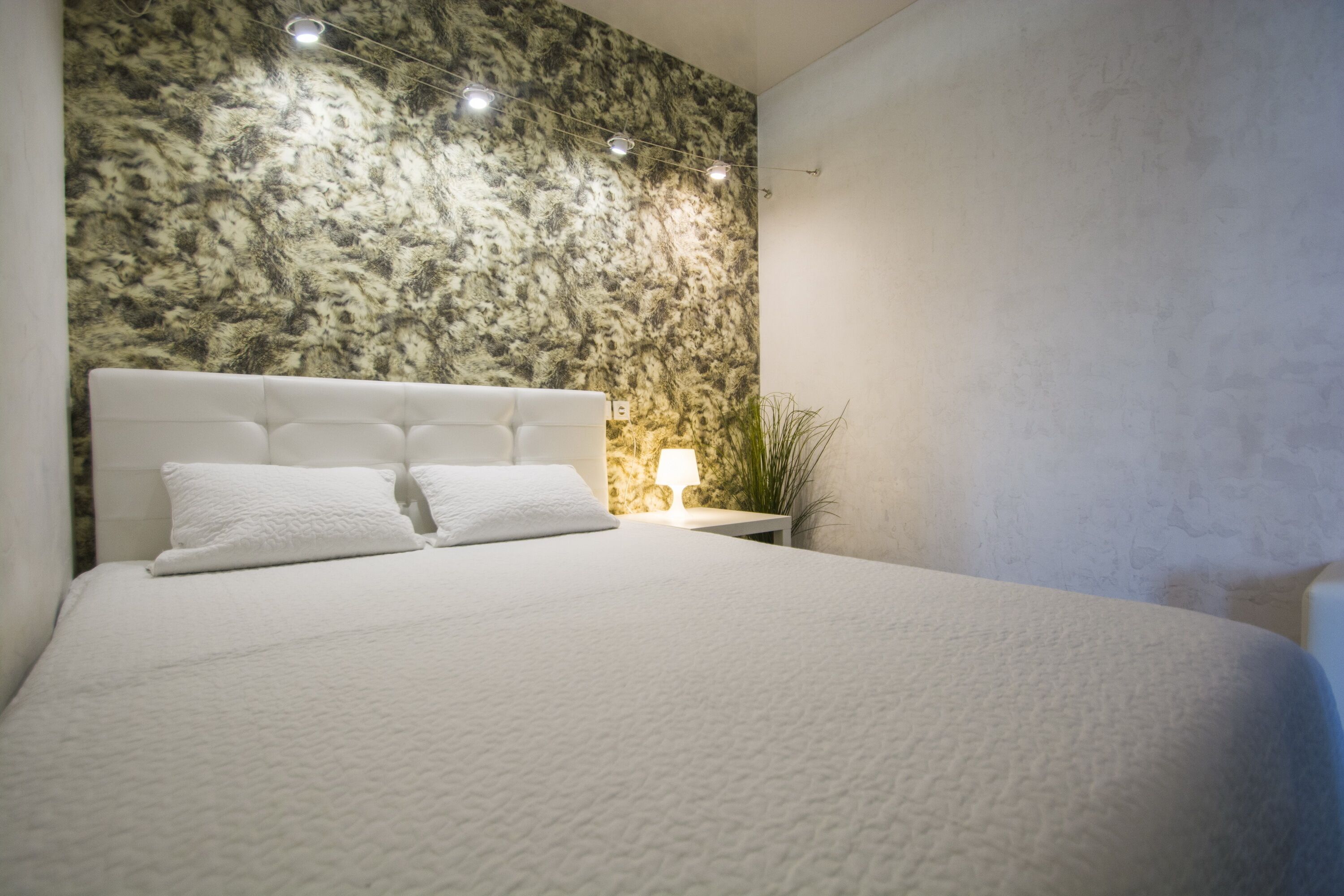 Ein ordentlich bezogenes Bett mit weißer Bettwäsche und Kissen, eine Wand mit Marmoroptik dahinter und ein kleiner Nachttisch mit Lampe und Pflanze.