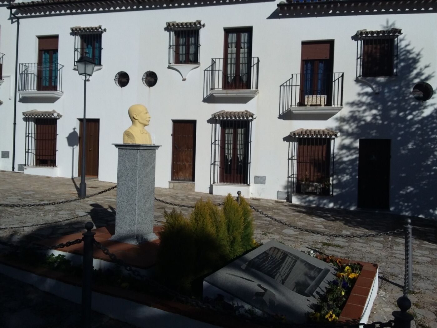 A courtyard with a bust sculpture, white buildings with balconies, and a small garden area.