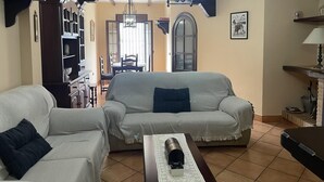 A living room with a sofa, armchair, and a coffee table. There is a bookshelf, a dining table with chairs, and a chandelier. - Grazalema - La Tassone: Charming house, fireplace and terrace (Grazalema)
