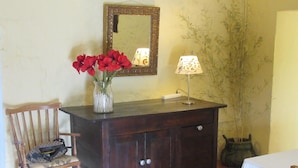 Ein hölzerner Kommode mit Spiegel darüber, eine Vase mit roten Blumen, eine Lampe und ein Stuhl mit einer Tasche darauf.