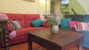 Ein gemütliches Wohnzimmer mit einem roten Sofa, einem Holztisch und einer Blumenvase. Im Hintergrund ist eine Treppe mit grünen Geländern zu sehen.