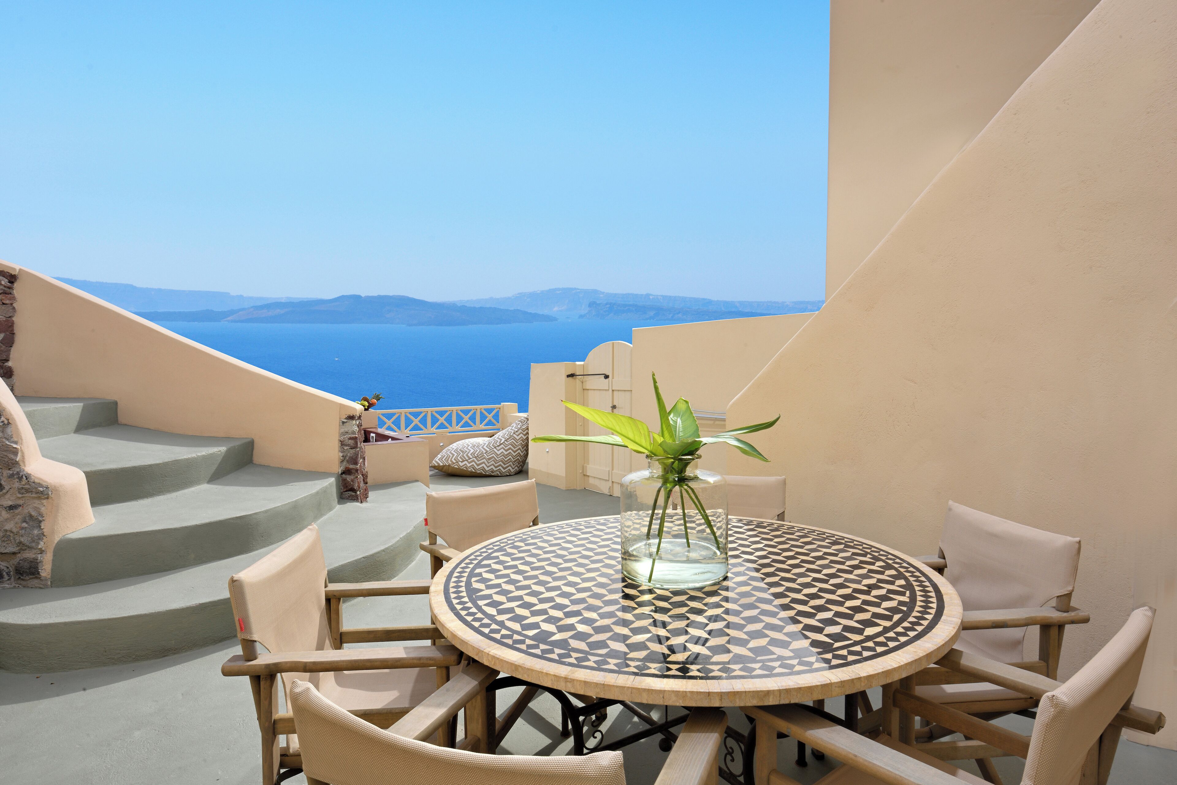 A round table with a checkered tablecloth, surrounded by wicker chairs, set on a balcony with a view of the sea and distant mountains.