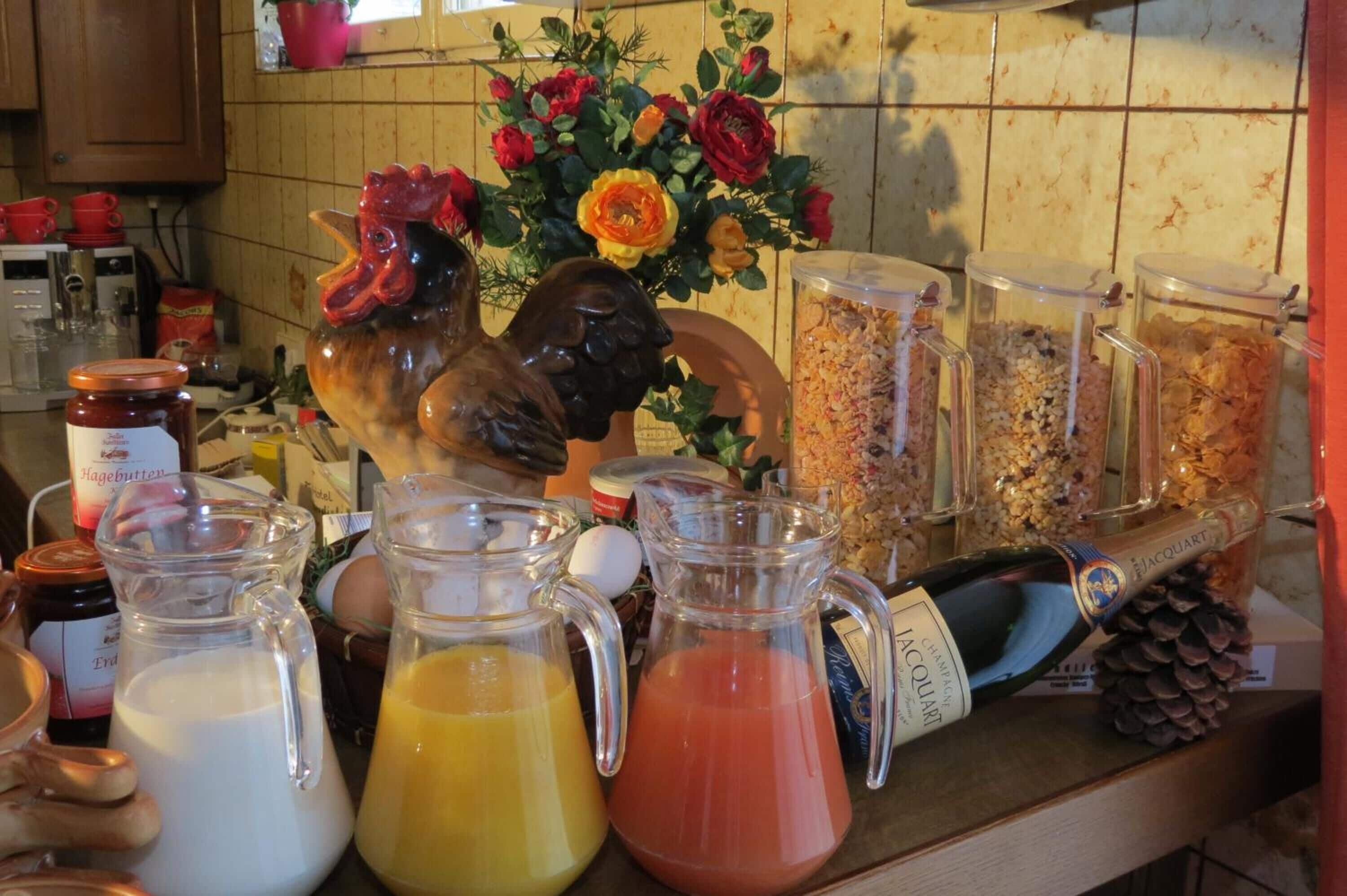 Uma bancada de cozinha com diversos objetos, incluindo uma figurineha de galo, arranjos de flores e recipientes com comida e bebidas.