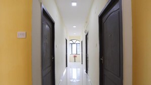 A hallway with two doors on each side, a light switch on the left wall, and a window at the end with a vase of flowers. - Green Tree Hotel (Chennai)