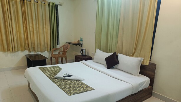 A neatly made bed with a white comforter and pillows, a small bedside table with a kettle, a chair, and a bottle of water on the wall. - Green Tree Hotel (Chennai)