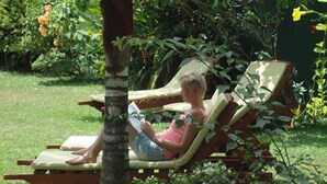 Una persona descansando en una silla de madera en un jardín con un árbol y follaje.