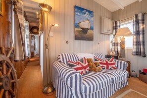 Une pièce avec un canapé à rayures, une lampe, un tableau représentant un bateau et une roue en bois.