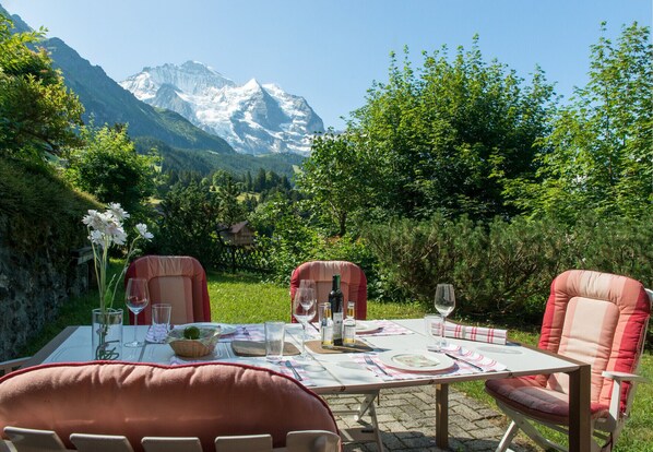 Mesa de comedor exterior con sillas, con montañas y follaje en el fondo.