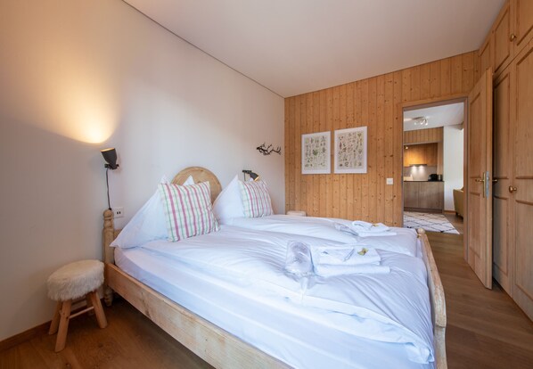 A bedroom with a wooden bed, a small stool, a wall-mounted lamp, and a door leading to a kitchen. - Chalet Sonneck - stylish apartment with garden (Wengen)