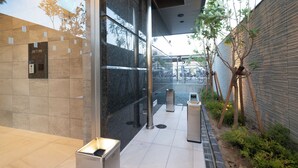 Un edificio moderno con fachada de vidrio, un poste de metal y un pequeño jardín con plantas y un árbol.