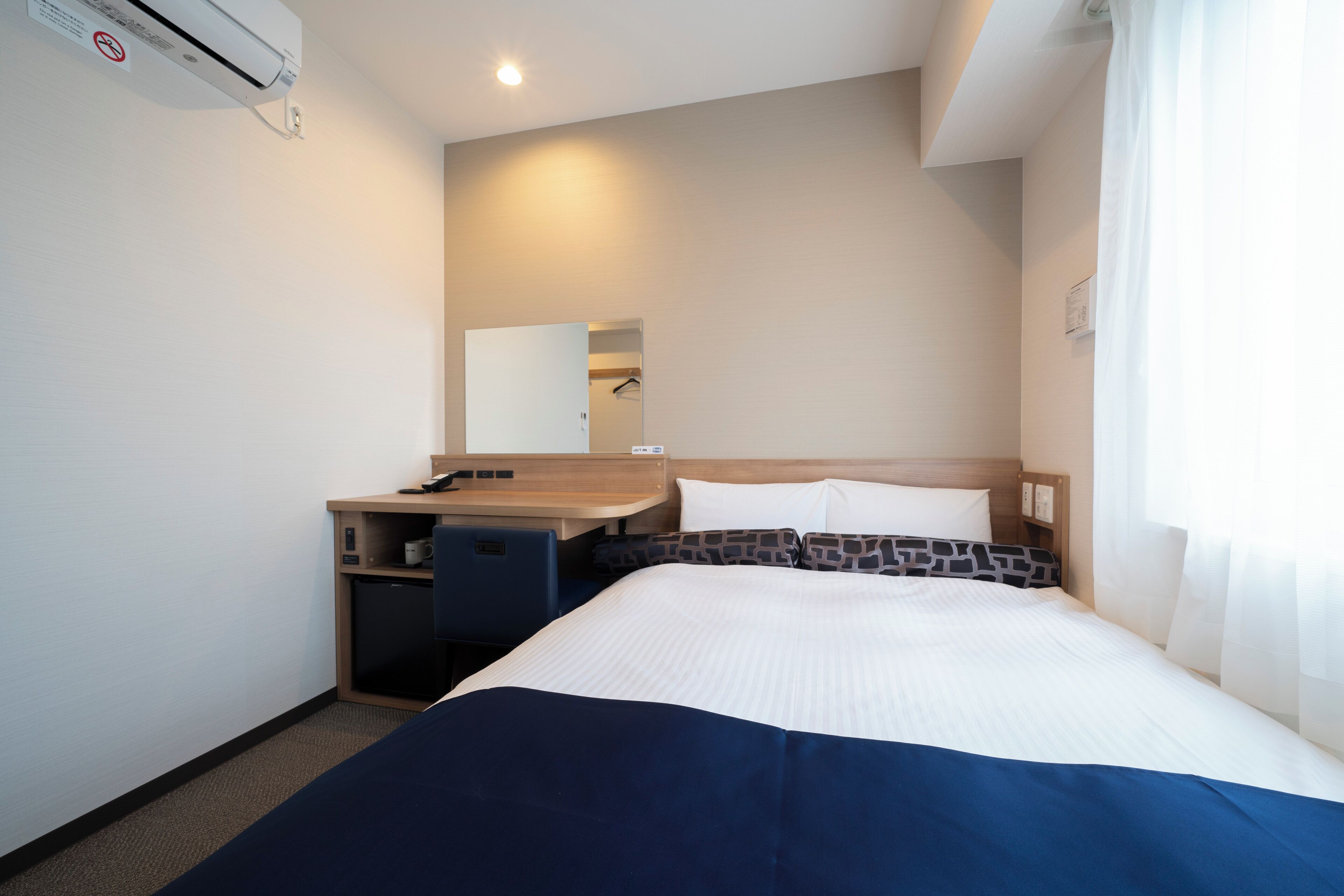 Una habitación de hotel con cama, escritorio con silla, refrigerador pequeño y espejo en la pared.