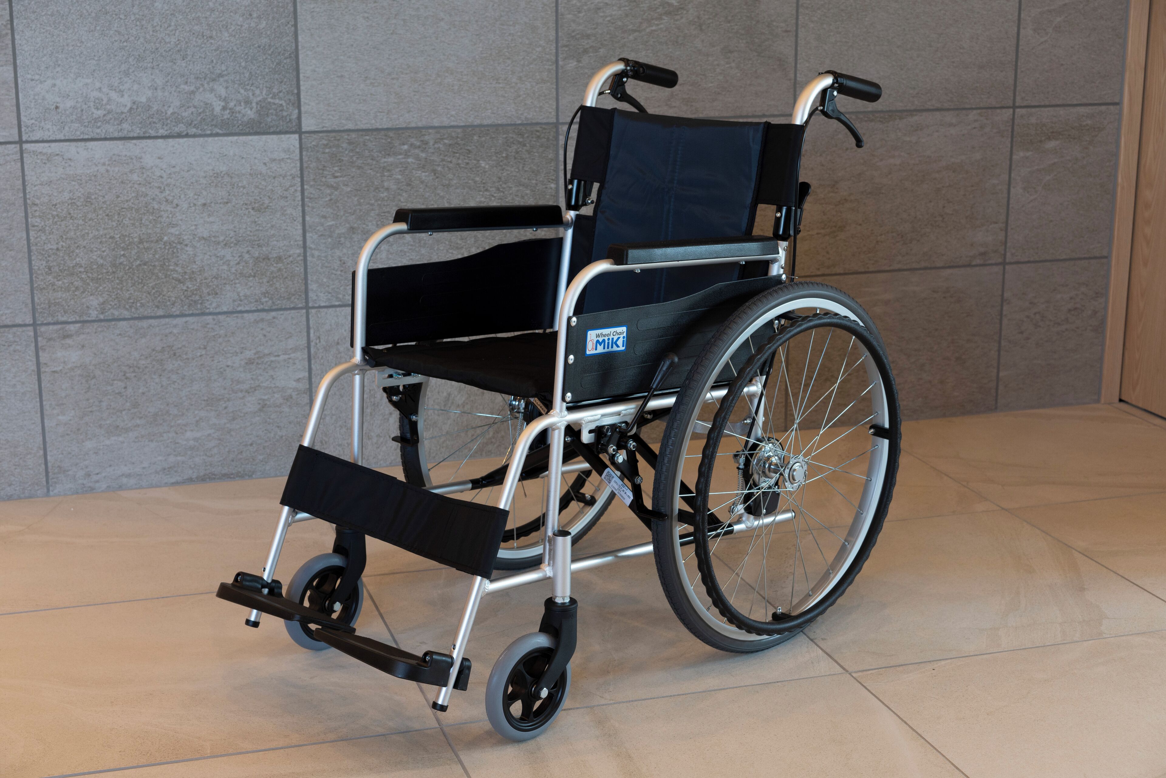 A wheelchair with a black seat and backrest, silver frame, and black wheels, positioned on a tiled floor against a grey wall.