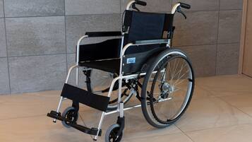 A wheelchair with a black seat and backrest, silver frame, and black wheels, positioned on a tiled floor against a grey wall.