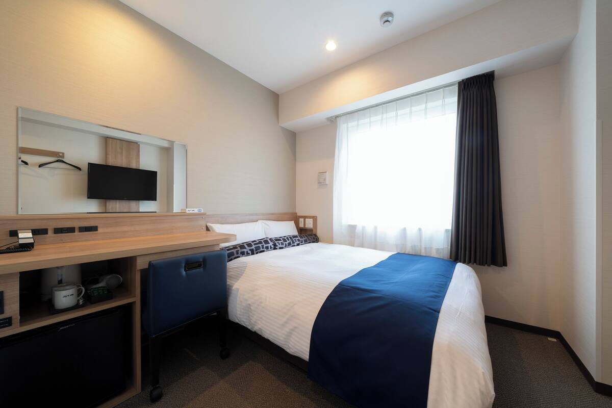 Quarto de hotel com cama, escrivaninha com computador, televisão e janela com cortinas.