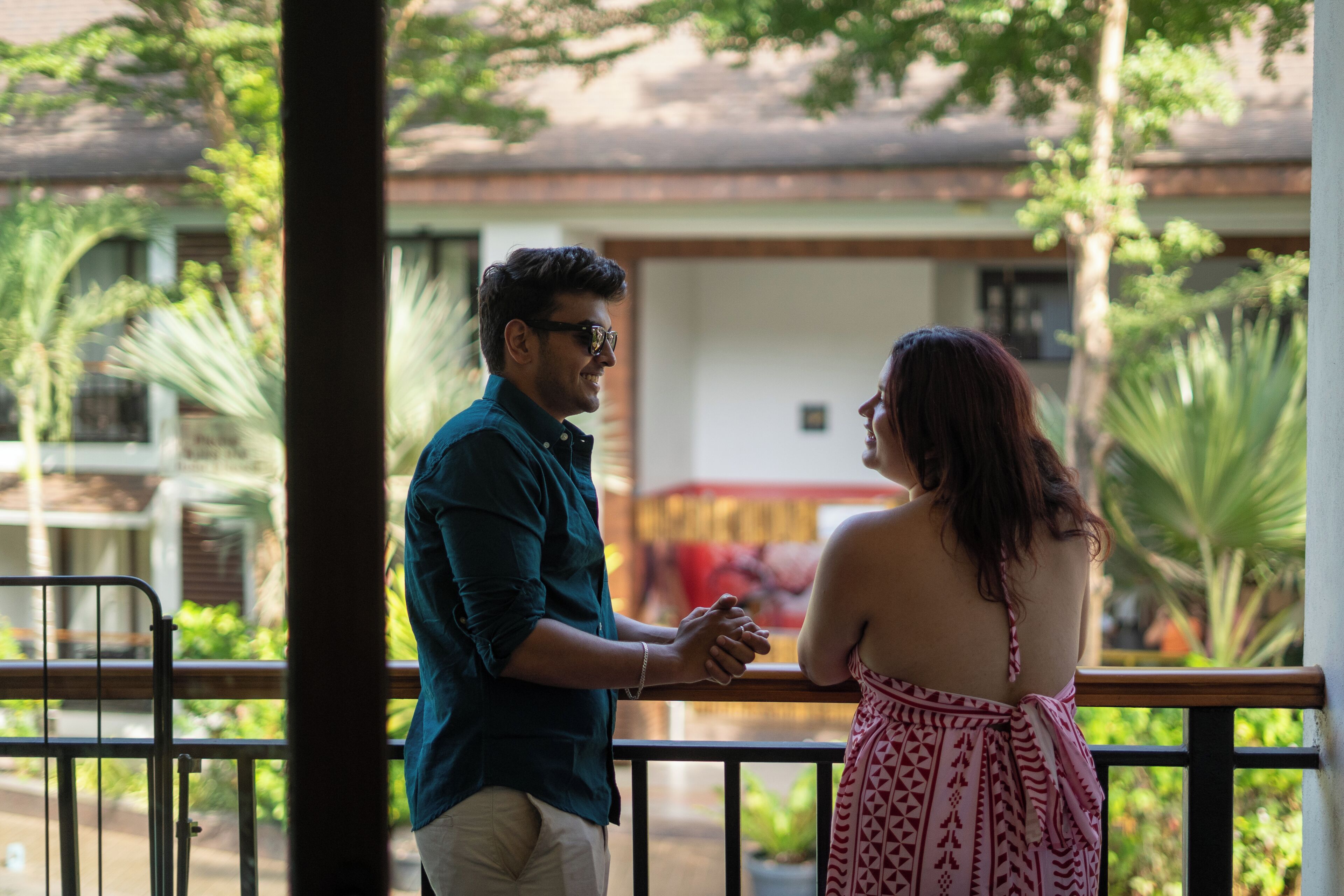 A man and a woman standing on a balcony with a railing, overlooking a building and greenery.
