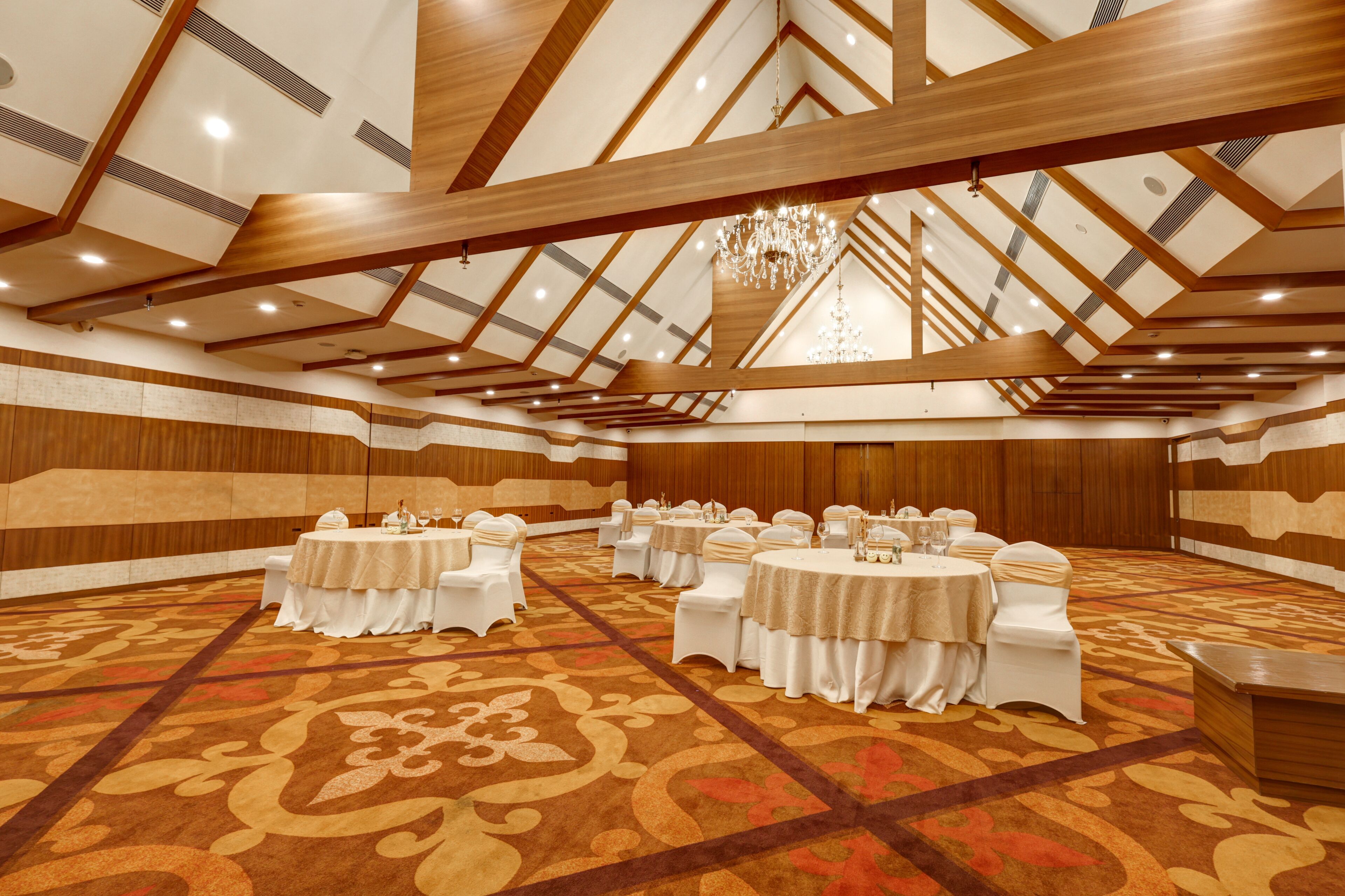 A large conference room with high ceilings, wooden beams, and a patterned carpet. Tables are set with white chairs and tablecloths.