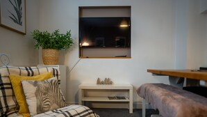 Una habitación acogedora con un sofá a cuadros, un banco, un estante blanco pequeño con un letrero que dice 'hello', una cesta trenzada con plantas verdes y un televisor montado en la pared.