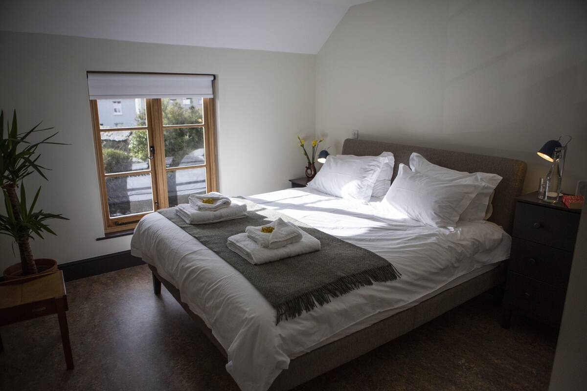Un lit bien fait, avec du linge de lit blanc, un plaid gris et deux serviettes posées sur une table de chevet en bois. Il y a une fenêtre avec un cadre en bois et une plante en pot sur la table de chevet.