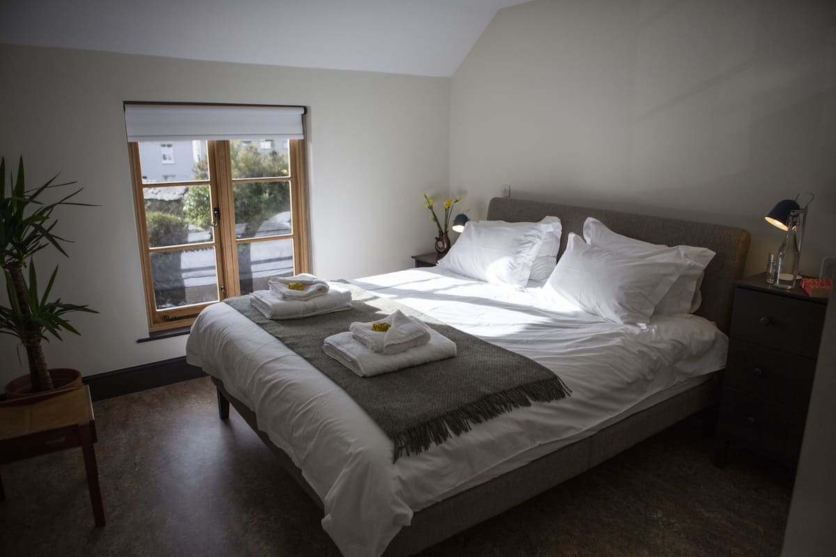 Un lit bien fait, avec du linge de lit blanc et un plaid gris, une table de chevet avec une lampe, et une fenêtre donnant sur les bâtiments environnants.
